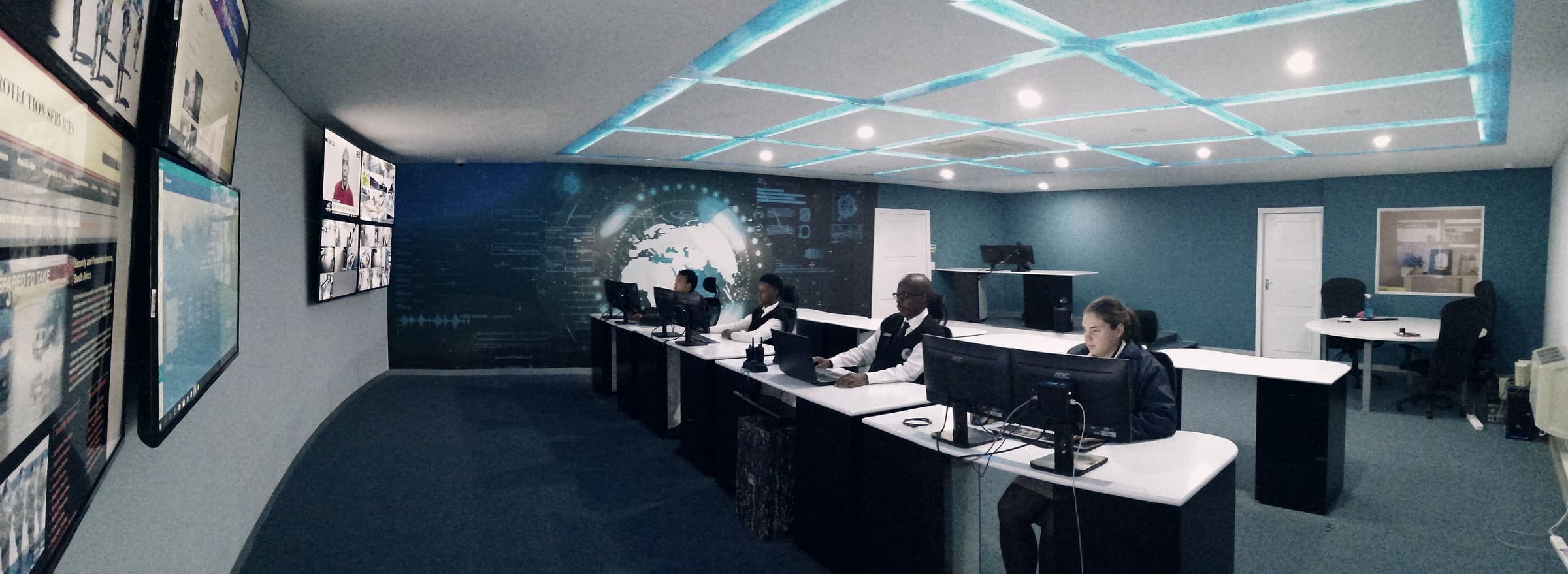 Wide panorama of control room: LED ceiling, video walls, four operators (phone panorama, slight distortion at edges)