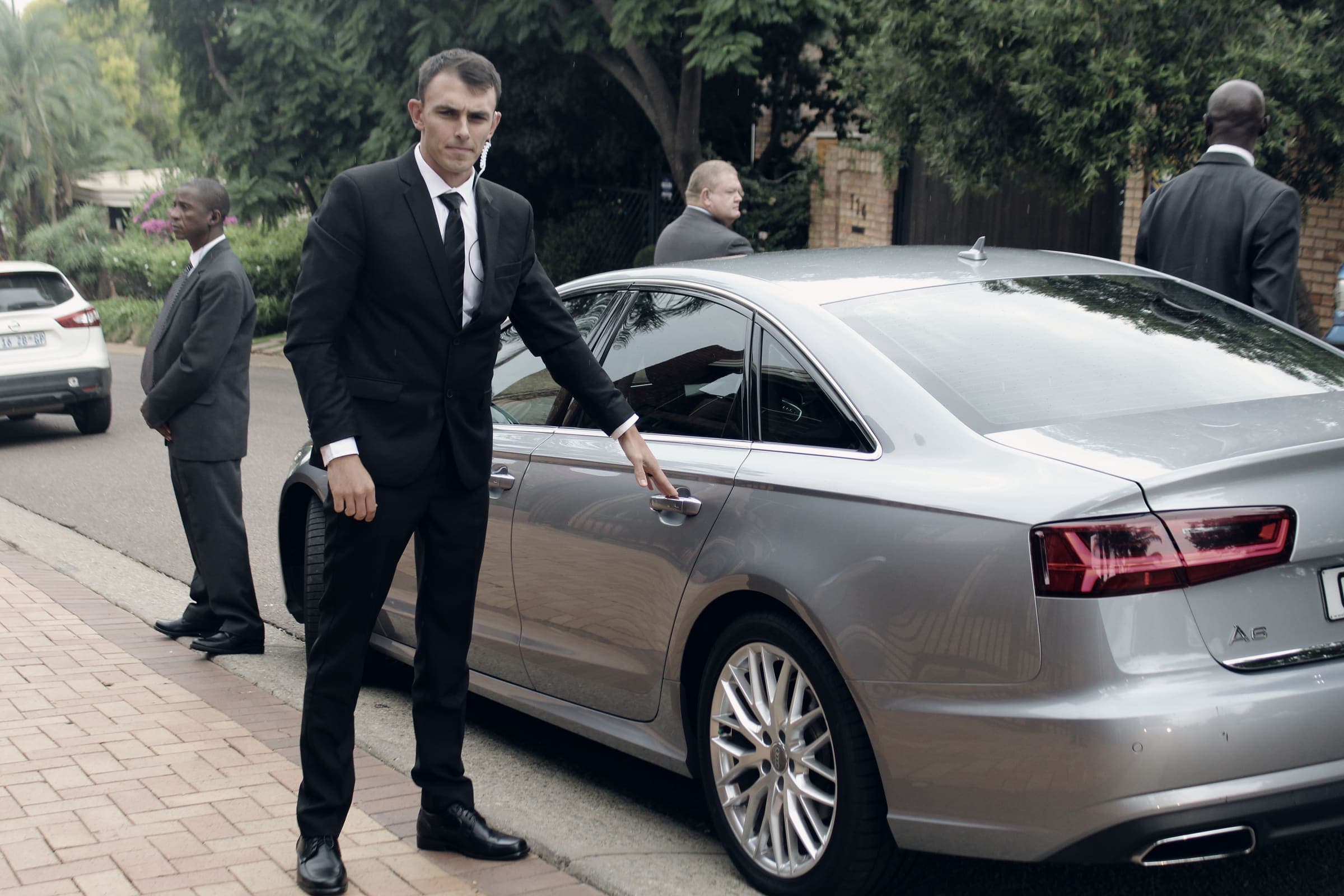 Suited close-protection officer with earpiece at the door of a silver Audi A6, team behind