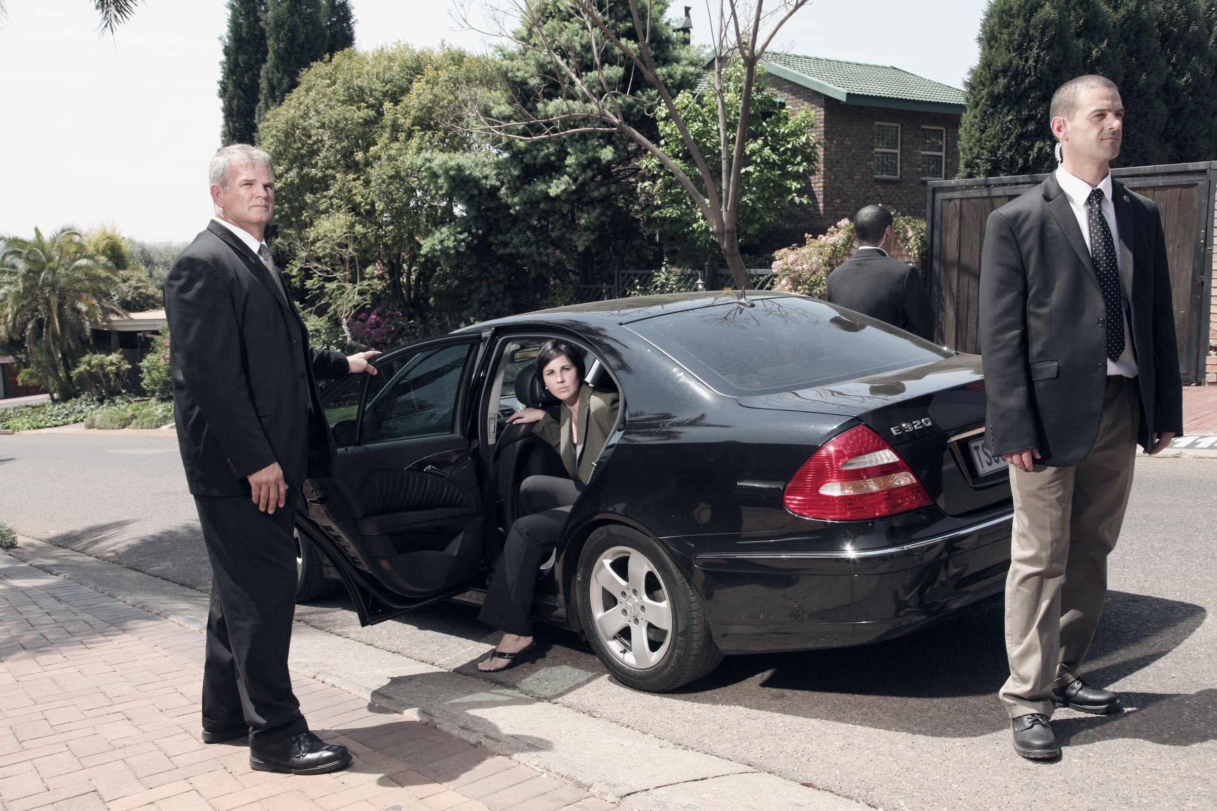 Two suited CPOs at the open rear door of a black Mercedes E-Class, principal seated, residential driveway