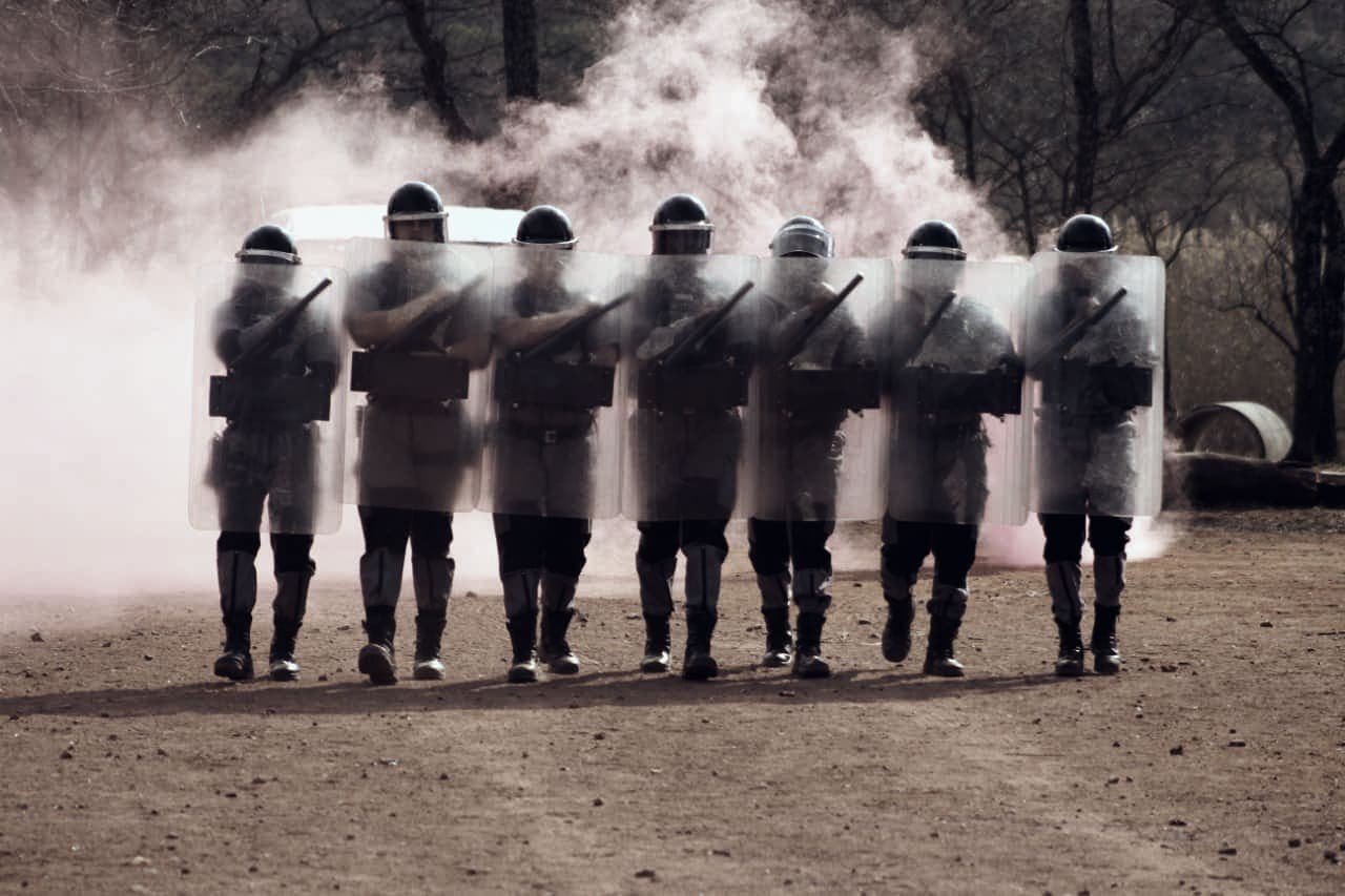 Riot shield line advancing through pink smoke