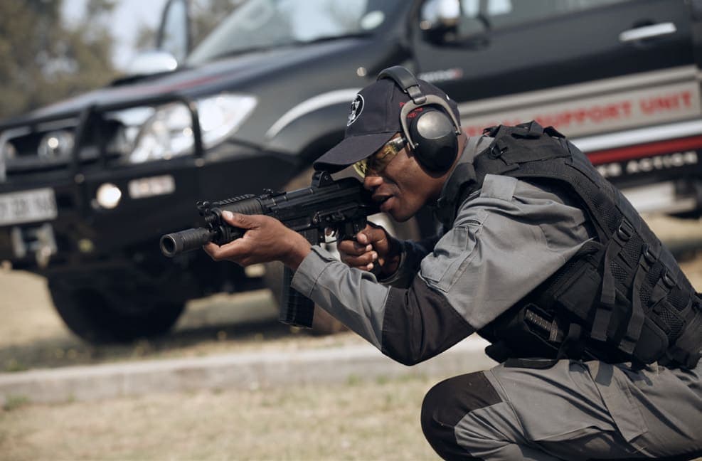 Officer with ear defenders aiming rifle beside Support Unit vehicle